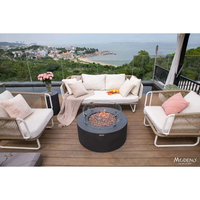 small round fire put table lit on lava rocks with round glass windscreen in between outdoor sofa with matching tank cover that is being used as a table top to hold a vase of flower