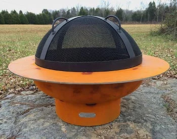 A round, orange fire pit with a black metal spark guard on top. The fire pit is placed on a rock in a grassy field.