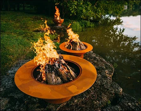 Two wood burning fire pits, one large and one small, both with flames burning inside. The larger fire pit is placed on a rock near a body of water.