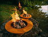 Two wood burning fire pits, one large and one small, both with flames burning inside. The larger fire pit is placed on a rock near a body of water.