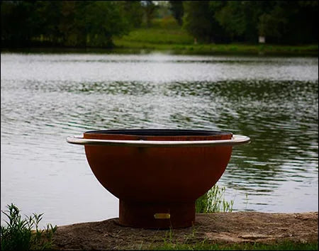 A round, rust-colored wood burning fire pit with a stainless steel handle. It is sitting on a rock near a lake with smoke rising from it.