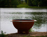 A round, rust-colored wood burning fire pit with a stainless steel handle. It is sitting on a rock near a lake with smoke rising from it.