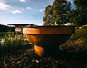  A round, rust-colored fire pit with a hexagonal rim. It is sitting on a rock near a body of water.