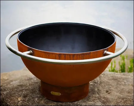 A round, rust-colored wood burning fire pit with a stainless steel handle. It is sitting on a rock with a dark interior.