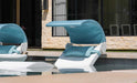 Two white in-pool loungers with blue canopies and cushions partially submerged in a pool, with a stone wall and building in the background.