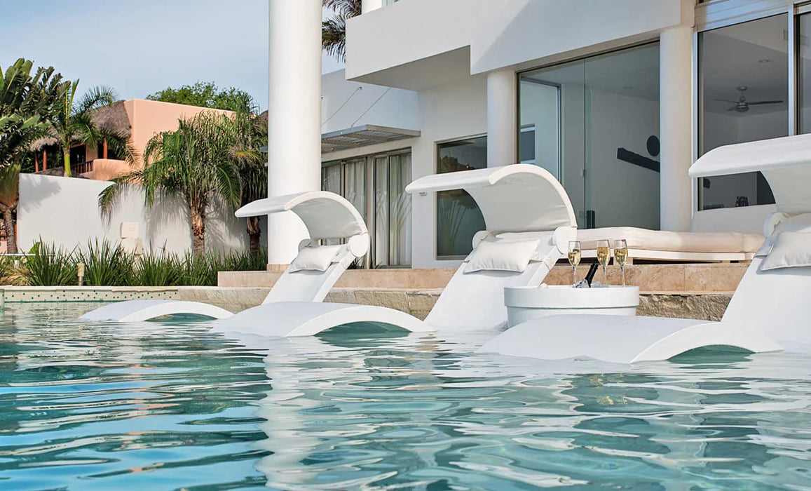 Three white in-pool loungers with white sun shades and cushions, plus a small table with drinks, partially submerged in a pool with a building and palm trees in the background.