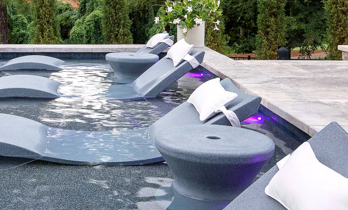 A luxurious poolside setup featuring multiple gray Ledge Lounger Signature Chaises with integrated tables. The chaises are floating in the water and each has a white pillow.