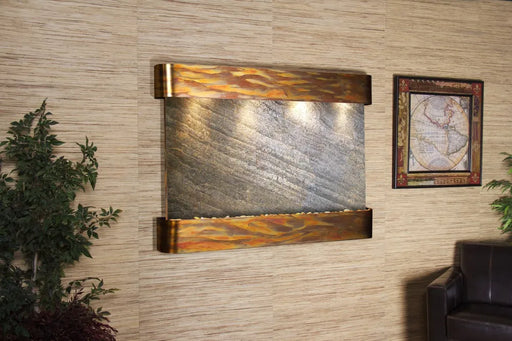 
A horizontal, rectangular wall-mounted water fountain featuring a textured natural stone panel with prominent diagonal striations in shades of grey and hints of brown. The top and bottom frames are made of a warm-toned, hammered or distressed metal, possibly copper, with a wavy, flame-like pattern and reflections of light. 