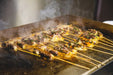 A close-up of a flat-top grill cooking skewers of meat. The meat is browned and sizzling, with steam rising from the grill.