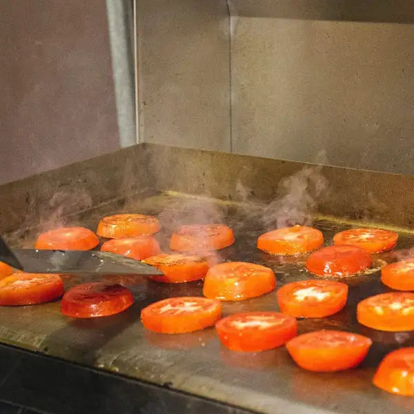 A close-up of a flat-top grill with a spatula turning over slices of tomato. The tomatoes are sizzling and releasing steam.