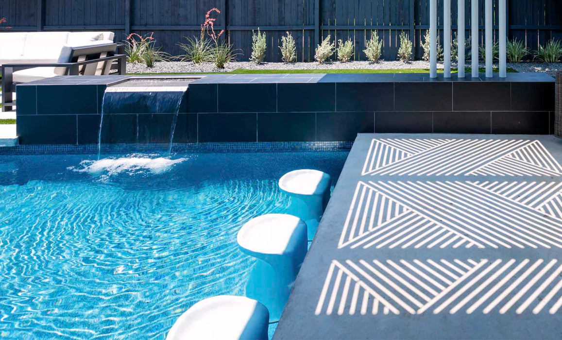 A modern poolside area with two white bar stools partially submerged in the water. A small waterfall cascades into the pool, and there is a geometric pattern cast on the pool deck.