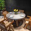 A round, light grey dining table is set for four people with white plates, silverware, and a vase of yellow flowers in the middle. The table is surrounded by four wooden chairs with woven seats and backs.