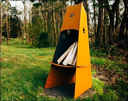 A tall, conical wood-burning fire pit outdoors with firewood inside. The fire pit is surrounded by green grass and trees.