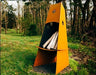 A tall, conical wood-burning fire pit outdoors with firewood inside. The fire pit is surrounded by green grass and trees.