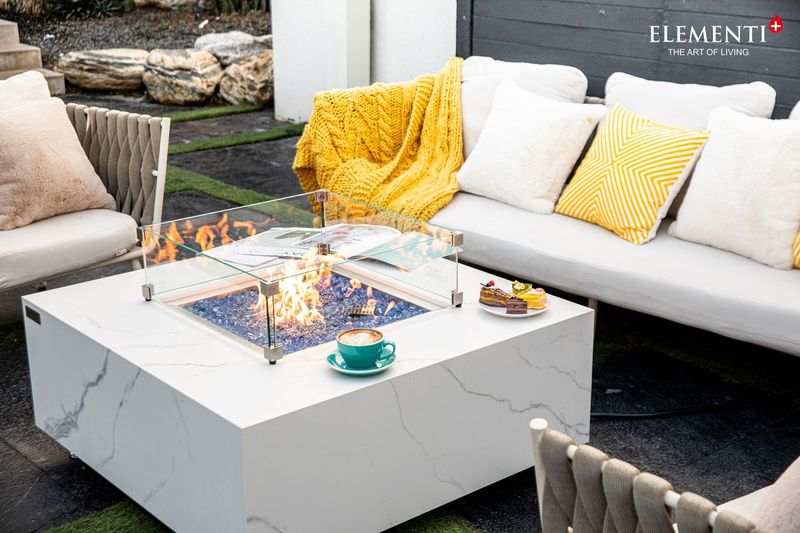 white marble fire table with protective glass and fire lit in the middle surrounded by protective clear glass. the fire table has plates of cakes and coffee on it