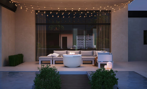 White Ledge Lounger Halo outdoor propane Fire Pit lit on a contemporary patio with sectional seating, string lights, and a pool in the background.