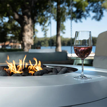 Close-up of a lit fire pit with a glass of rosé wine resting on the edge, with blurred trees and a lake in the background.