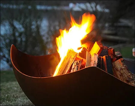 A close-up of a fire pit with flames licking at the burning wood inside. The fire pit has a unique, manta ray-shaped design.