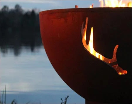 Wood burning fire pit with a deer antler cutout design. The fire pit is round and made of rusted metal. Flames are burning brightly inside, illuminating the deer antler silhouette. The fire pit is placed outdoors near a lake at dusk. The sky is a soft blue with hints of pink and purple.