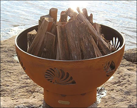 A round, rusted metal fire pit filled with firewood. The fire pit has seashell cutouts and is placed on a rock near a body of water. The sun is shining on the fire pit.