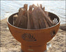 A round, rusted metal fire pit filled with firewood. The fire pit has seashell cutouts and is placed on a rock near a body of water. The sun is shining on the fire pit.