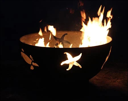 A round, rusted metal fire pit with flames burning inside. The fire pit has a seahorse and starfish design cut into the metal. The background is dark.