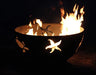 A round, rusted metal fire pit with flames burning inside. The fire pit has a seahorse and starfish design cut into the metal. The background is dark.