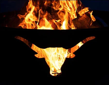 A close-up image of a wood-burning fire pit with a longhorn cutout design. The fire is burning brightly, illuminating the cutout.