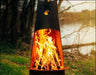  A tall, conical wood-burning fire pit with flames burning inside. The fire pit is surrounded by green grass and is near a body of water. There is a deer logo on the side of the fire pit.