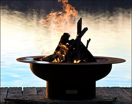 Wood burning fire pit with a roaring fire. The fire pit is round and made of rusted metal. The flames are orange and yellow and are rising high into the air. The fire pit is placed on a wooden dock by a lake.