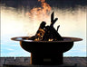 Wood burning fire pit with a roaring fire. The fire pit is round and made of rusted metal. The flames are orange and yellow and are rising high into the air. The fire pit is placed on a wooden dock by a lake.