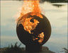 A spherical fire pit with a map of the Earth etched into its surface. Flames are rising from the center of the fire pit, illuminating the continents. The fire pit is placed on a rock near a body of water.