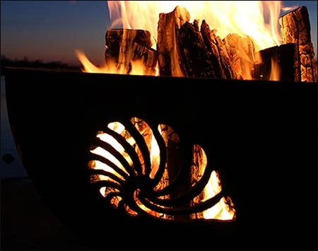 A close-up of a wood-burning fire pit at night. The fire pit has a seashell design cut into the metal. Flames are visible through the cutout.