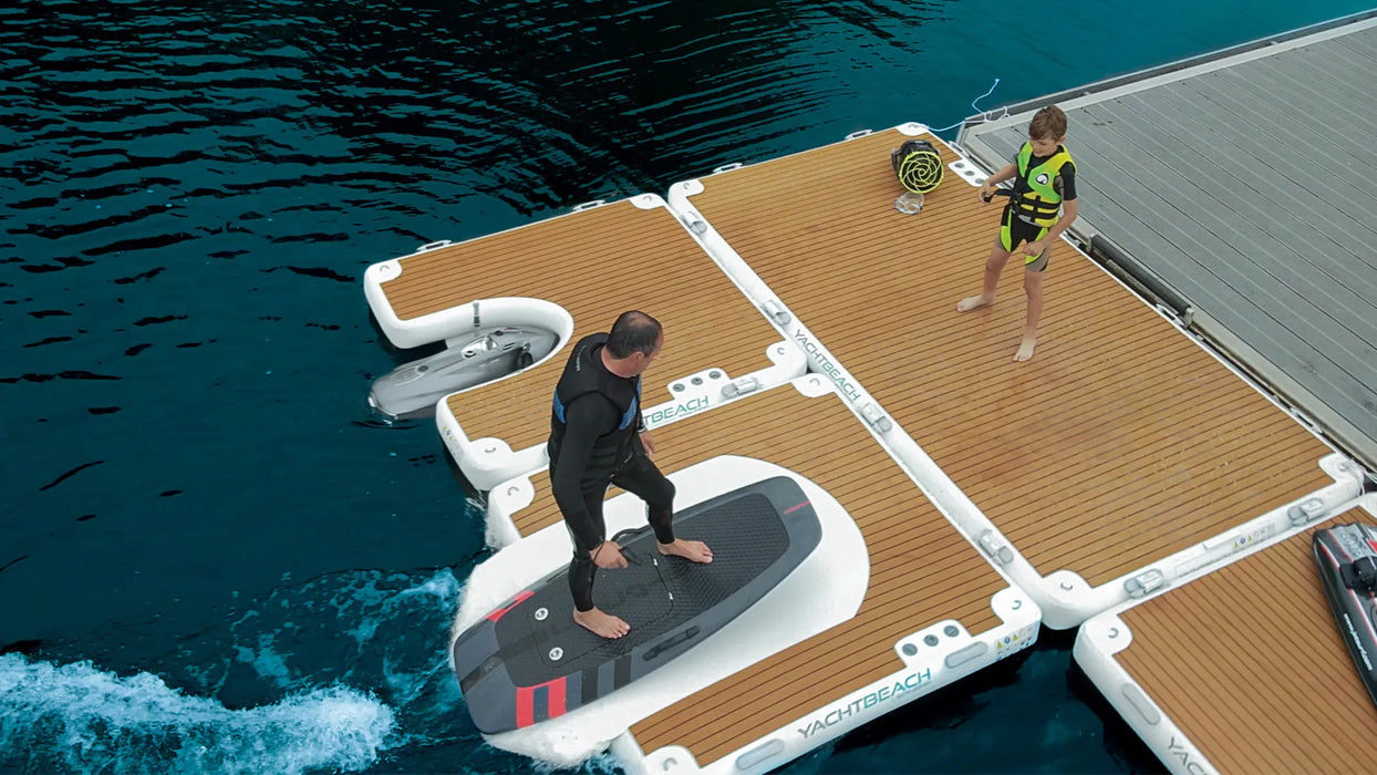 Two people on a floating dock with a surfboard, surrounded by water.