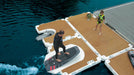 Two people on a floating dock with a surfboard, surrounded by water.
