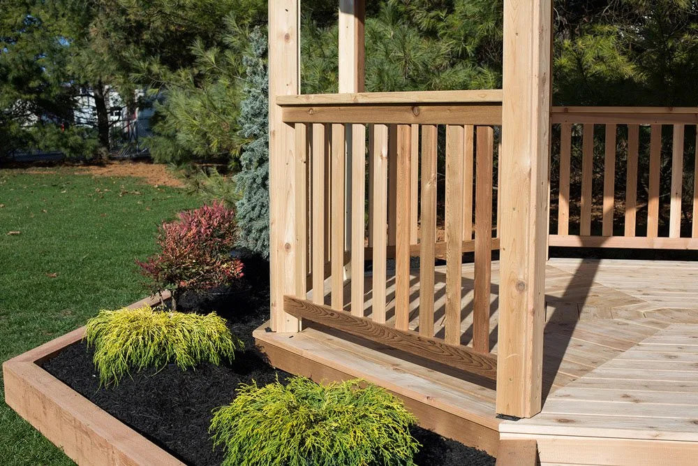 A wooden octagonal gazebo with a shingled roof. The gazebo is surrounded by lush greenery and features a patio furniture set inside.